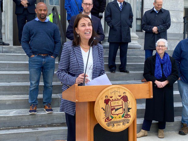 Woman at wooden podium outdoors in front of courthouse steps with spectators behind her.