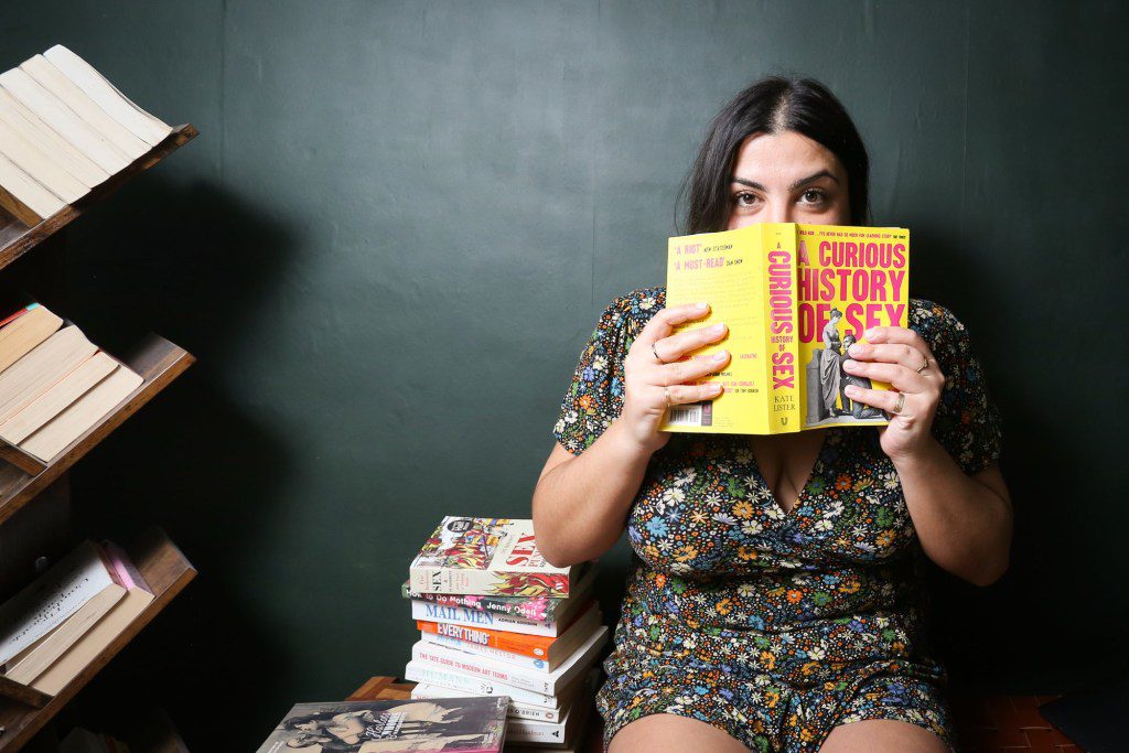 Almara Abgarian holding a book ('A Curious History of Sex') in front of her face, with other books piled up next to her/on shelves to her right