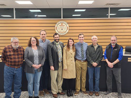 8 people standing in front a meeting table with a township seal on the wall in the background